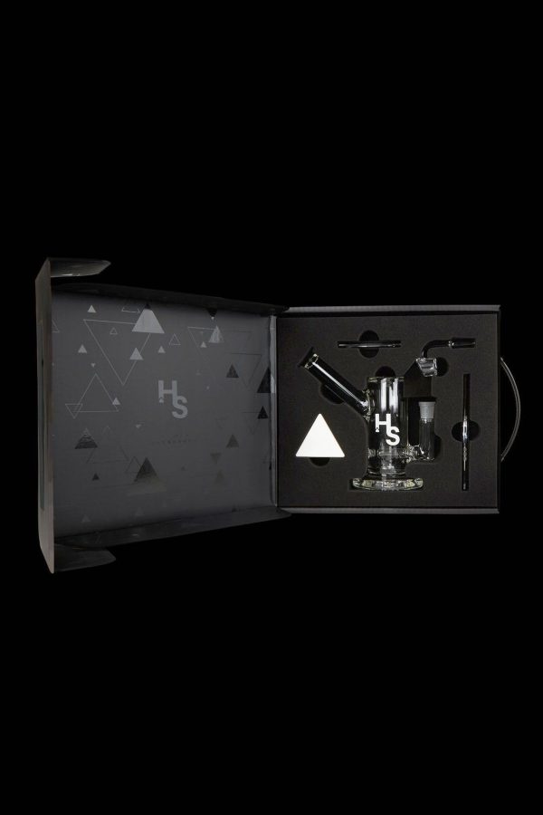 Open black gift box displaying cannabis smoking accessories, including a glass pipe, vaporizer, and tools, with a geometric pattern and "Hs" logo.