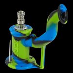 Green and blue silicone oil rig with a metal bowl and dab tool, isolated on a black background.
