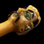 Hand-blown glass cannabis pipe sculpted as a grotesque face with a single green eye and swirling amber details.