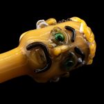 Hand-blown glass pipe shaped like a cartoon character's head, featuring green eyes and a yellow and black color scheme, against a black background.