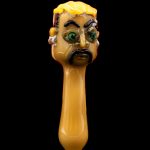 Hand-blown glass pipe shaped like a cartoonish face with green eyes, against a black background.
