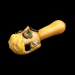 Hand-blown glass pipe sculpted as a grotesque, smiling face with green eyes, against a black background.