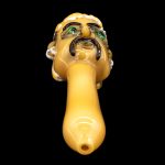 Yellow glass pipe sculpted as a grotesque face with green eyes, studio shot on black background.