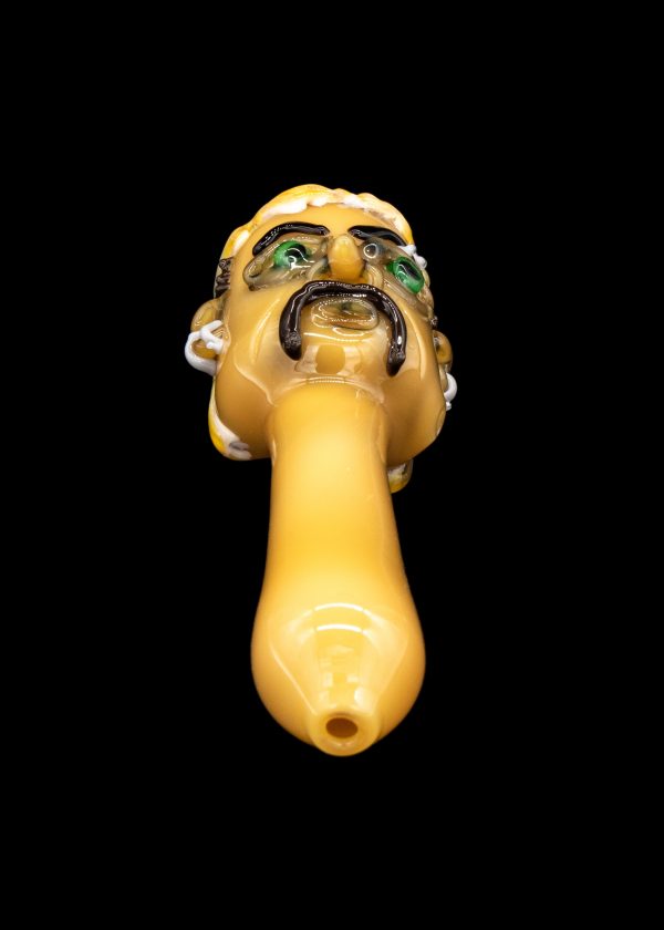 Yellow glass pipe sculpted as a grotesque face with green eyes, studio shot on black background.