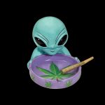 Ceramic ashtray shaped like an alien with a lit joint resting on a cannabis leaf design.