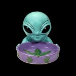 Turquoise alien head ashtray with cannabis leaf design on a purple base, isolated on black.