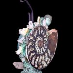 Intricate, handmade glass water pipe shaped like an ammonite fossil, adorned with quartz crystals and teal-colored detailing, set against a black background.