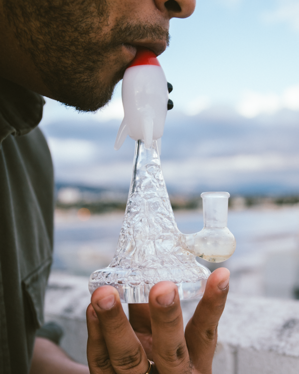 A person holds and inhales from a rocket-shaped glass water pipe, with a blurred landscape background.