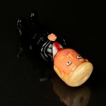 Glass sculpted cannabis bud with a comical, cartoonish face, resting on a black glass surface with a reflective sheen.