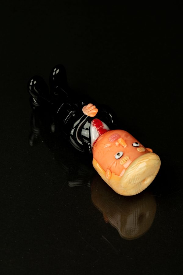 Glass sculpted cannabis bud with a comical, cartoonish face, resting on a black glass surface with a reflective sheen.