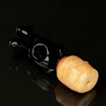 Glossy black glass pipe resembling a hand, with a peach-colored mouthpiece, isolated on a black reflective surface.