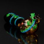 Hand-blown glass cannabis pipe with intricate amber, turquoise, and green floral detailing, displayed on a reflective black surface.