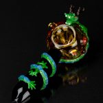 Hand-blown glass pipe with a vibrant green lizard design and intricate detailing, displayed on a reflective black surface.