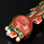 Close-up of a colorful, hand-blown glass cannabis pipe with a bulbous, reddish-orange chamber and intricate green and white details, reflecting on a dark surface.