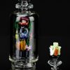 Hand-blown glass water pipe featuring colorful insect and mushroom designs, with a separate glass piece alongside, all reflected on a dark surface.