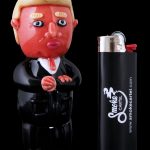 A sculpted Donald Trump figurine stands beside a black Smoke Cartel lighter, both against a dark background.