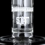 Close-up of a clear glass water pipe with “STAX” etched on the side, filled with water and reflecting on a dark surface.