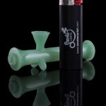 Green glass dab rig and black lighter with cannabis-themed branding, reflected on a black surface.