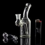 Clear glass water pipe (bong) with loose glass marbles and a lighter reflected on a glossy black surface.