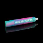 A vibrant pink and blue cannabis oil cartridge with “HUMAN SUCKS” printed on its transparent window, reflected on a black surface.