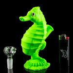 A vibrant green glass seahorse sculpture displayed alongside a glass smoking bowl and lighter, reflecting on a glossy black surface.