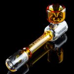 Glass cannabis pipe with amber and clear glass, reflected on a dark surface.