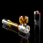 Glass cannabis pipe and lighter on a reflective black surface.