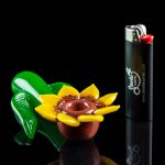 A vibrant green and yellow glass flower pipe rests beside a black lighter branded with "Smoke Cartel" on a reflective black surface.