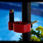Red cannabis accessory holder attached to a black pole, containing a lighter and rolled joint, with blurred green foliage and a blue background.