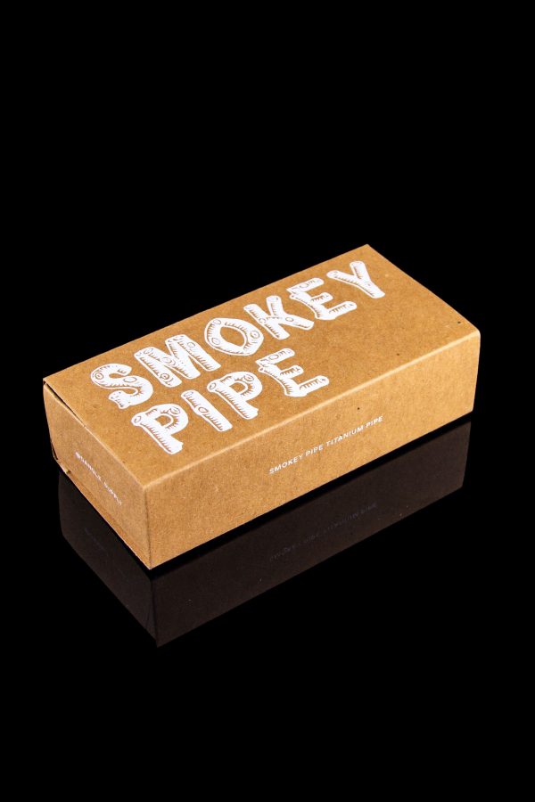 Cardboard box for “Smokey Pipe” titanium pipe, reflecting on a glossy black surface.