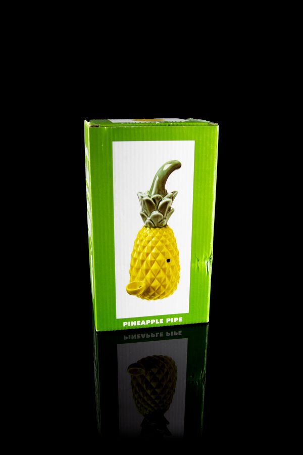 Retail packaging for a yellow ceramic pineapple-shaped cannabis pipe, reflected on a glossy surface.