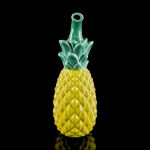 Yellow glass pineapple-shaped water pipe with a green leafy top, reflecting on a black surface.