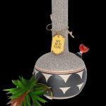 A patterned ceramic water pipe (“Bud Vase”) with a tag and flowering plant beside it, against a black background.