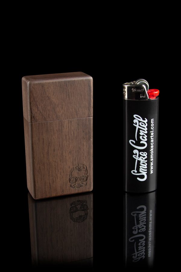 Dark wood cannabis storage box with engraved design next to a lighter branded with “Smoke Cartel” logo, reflected on a black surface.