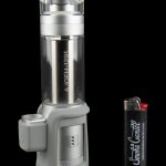 A silver and black electronic dab rig with a glass water pipe attachment, positioned next to a lighter with cannabis-related branding.