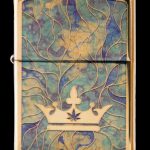 Gold Zippo lighter with a raised cannabis leaf within a golden crown design on a swirling teal and gold background.