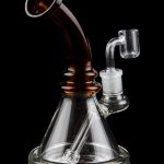 A clear glass water pipe (rig) with a colored mouthpiece and a quartz nail for vaporizing concentrates, isolated on a black background.