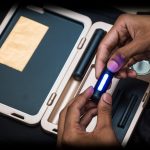 Hands holding a lit, glowing cannabis vaporizer pen over an open, wood-toned cannabis accessory case with rolled joints and a grinder inside.