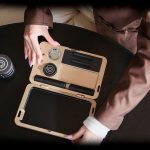 Overhead view of hands opening a sleek, gold cannabis accessory case containing various tools and compartments on a dark surface.