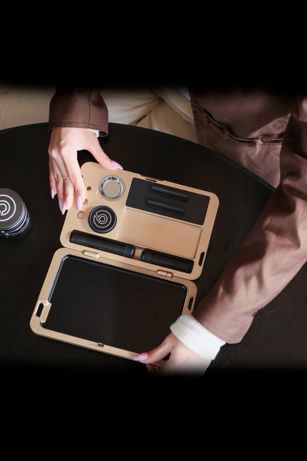 Overhead view of hands opening a sleek, gold cannabis accessory case containing various tools and compartments on a dark surface.