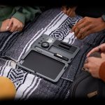 Overhead view of a cannabis storage case open on a patterned blanket, surrounded by people's hands and clothing.
