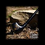 A black pipe with gold detailing rests on a tree stump surrounded by dry leaves and earth, evoking imagery from *The Lord of the Rings*.