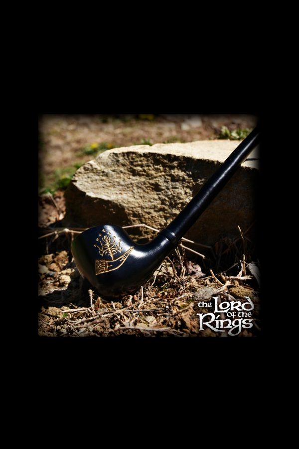 A black pipe with gold detailing rests on a tree stump surrounded by dry leaves and earth, evoking imagery from *The Lord of the Rings*.