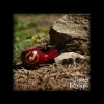 A red glass pipe with intricate gold detailing rests on a weathered tree stump amidst green grass and brown earth, evoking a natural, earthy aesthetic alongside “The Lord of the Rings” logo.
