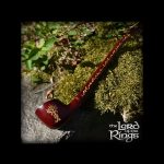 A long, intricately carved wooden pipe resembling a stylized branch lies on moss, with “The Lord of the Rings” logo visible below.