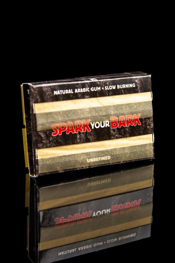 Stacked boxes of “Spark Your Bark” natural arabic gum slow-burning rolling paper, reflecting on a dark surface.