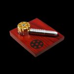 Gold and silver cannabis pipe and grinder on a red wooden tray with a circular logo.