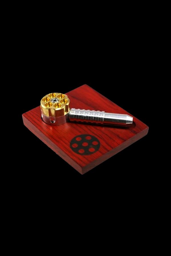 Gold and silver cannabis pipe and grinder on a red wooden tray with a circular logo.
