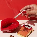 A hand with red nails holds a lit joint above an ashtray and matches, all set against a red and pink background.