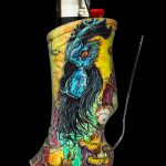 A colorful, patterned lighter sleeve depicting an ornate owl with a glass stirring rod beside it, against a black background.